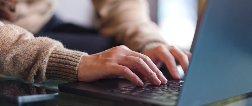 Close-up of hands typing on a laptop keyboard. The person is wearing a brown sweater. The setting appears relaxed, suggesting a casual work environment for Forex Trading Beginner.