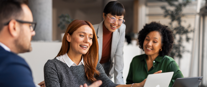 Four colleagues gathered around a laptop, engaged in a lively discussion about a Forex Trading Plan. They are smiling, conveying a collaborative and positive atmosphere.