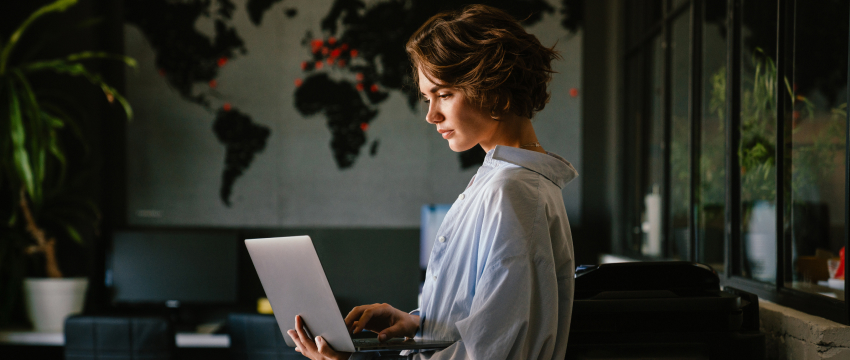 A woman in a light blue shirt stands in a modern office using a laptop, with a world map decorating the dark wall, creating a professional atmosphere focused on Forex Currency Strength and global market analysis.