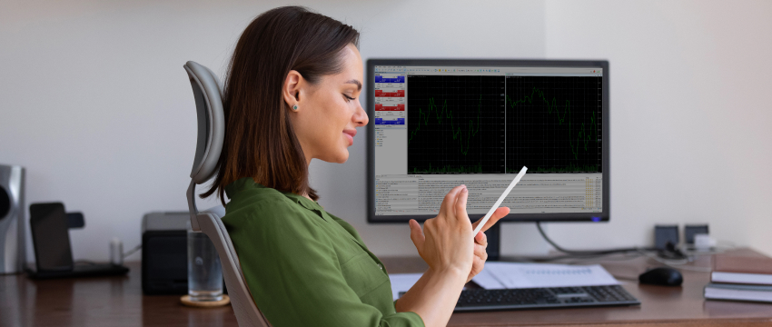 A woman in a green blouse sits at a desk using a tablet, with a computer monitor displaying stock graphs. The scene conveys focus and productivity, representing MetaTrader 5 for Beginners.