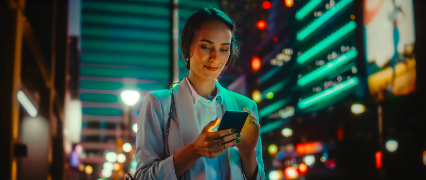 A woman in a white blazer smiles while using her smartphone at night on a bustling city street, illuminated by colorful neon lights and signs, representing Trading Sessions in Forex.