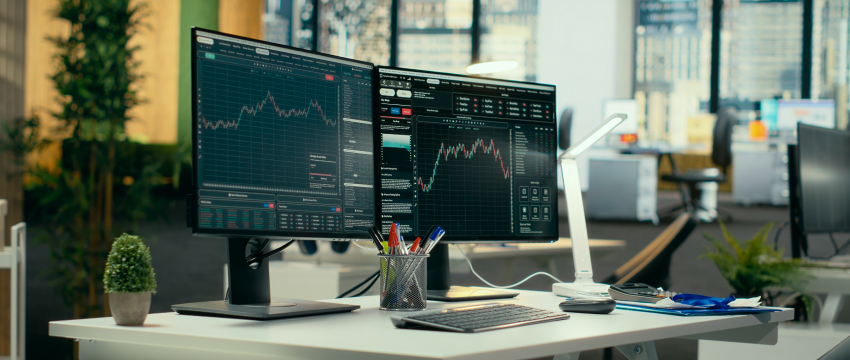 A modern office desk with two monitors displaying stock market graphs, highlighting algorithmic trading tools. The setup includes a keyboard, mouse, plant, pen holder, and documents, with a bright and professional ambiance.