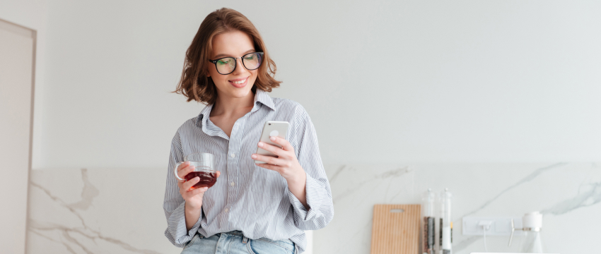 A woman with glasses smiles while looking at her smartphone in a modern kitchen, best crypto wallet for beginners. She holds a glass of red wine, creating a relaxed and cheerful atmosphere.