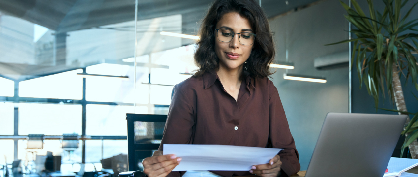 Businesswoman in a brown blouse and glasses reviews a document at her desk, with a laptop open, focusing on risk to reward ratio analysis. The modern office setting suggests focus and productivity.
