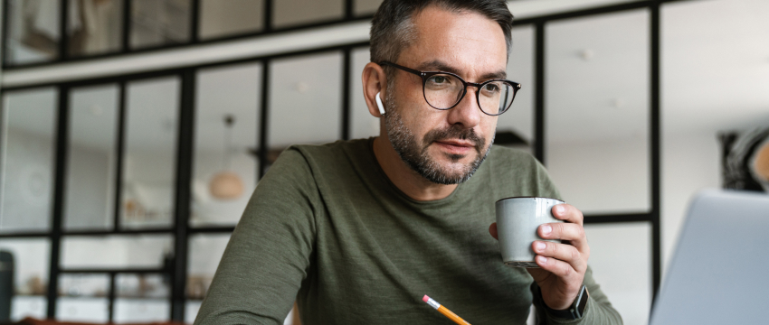 Man wearing glasses and earbuds works on a laptop at a modern desk while holding a mug and pencil, illustrating online trading education and what is CFD trading.