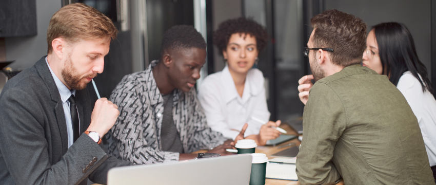 A diverse group of five people engaged in a serious business meeting around a table with laptops and coffee cups, conveying focus, collaboration, and a professional forex job environment.
