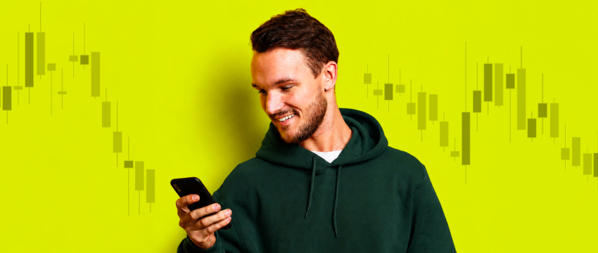 A man in a green hoodie looks at mobile market charts and smiles, set against a yellow background with candlestick graphs explaining what is FOMO in trading.