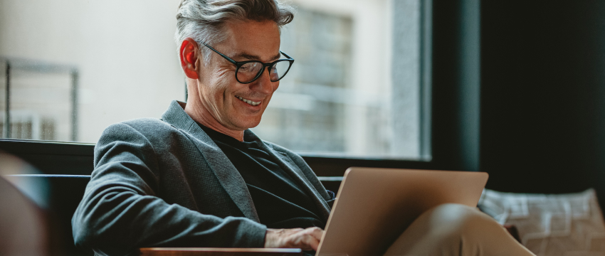 A man in a blazer and glasses smiles while looking at his laptop screen, depicting a user managing a Forex traders funding account.