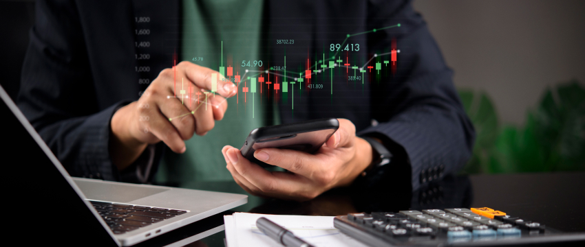 Trader analyzing financial market chart trends on a smartphone with a laptop, paper reports, and an Interest Rate Calculator on the desk.