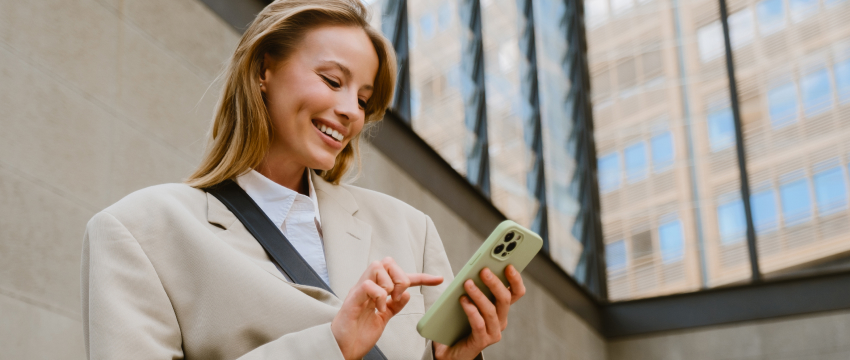A smiling businesswoman using a smartphone outside a modern office building, checking market updates and executing a support and resistance trading strategy on a mobile app.