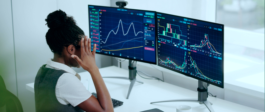 A side-view of a female trader sitting at her desk holding her head in stress while looking at multiple monitors displaying volatile financial market charts, highlighting top trading psychology tips for managing fear and emotional decisions.