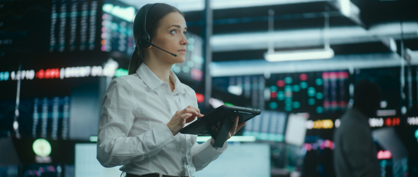 Trader wearing a headset reviewing market data on a tablet in front of live stock price screens